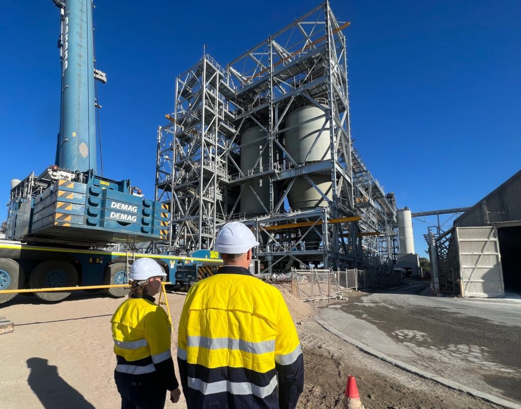 Two people wearing hard hats and long sleeve hi-vis PPE, standing outdoors, looking onto a industrial processing facility