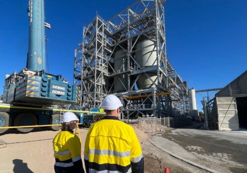 Two people wearing hard hats and long sleeve hi-vis PPE, standing outdoors, looking onto a industrial processing facility